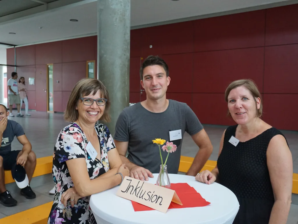 Drei Personen an einem Stehtisch mit dem Schild Inklusion.